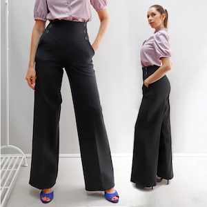 Può includere: Pantaloni neri a vita alta e gamba larga con dettagli di bottoni. I pantaloni hanno una vestibilità sartoriale e presentano una vita alta con dettagli di bottoni. La modella indossa i pantaloni con una camicetta lilla chiaro e scarpe blu.