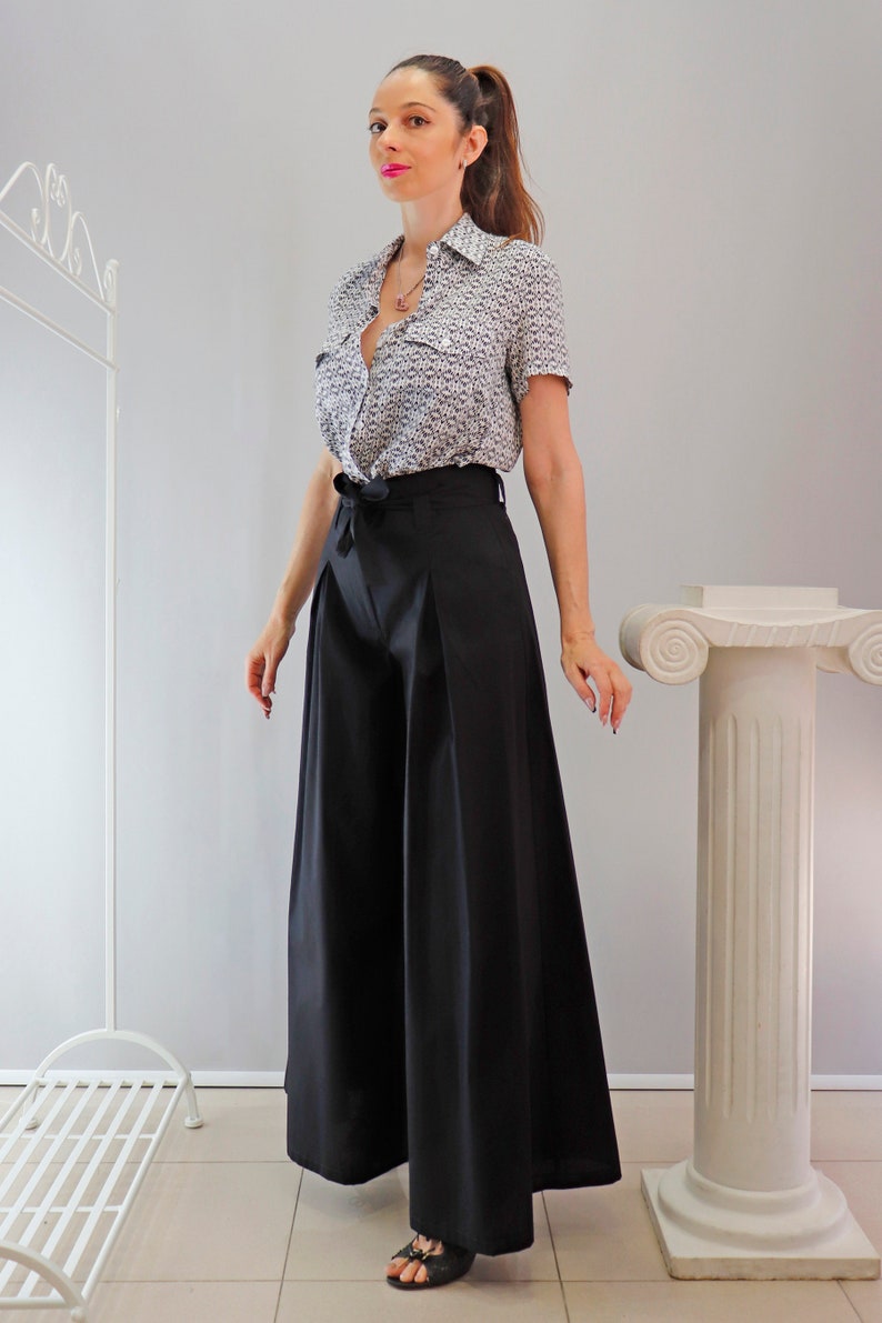 May include: A woman wearing a short-sleeved button-down shirt with a small, repeating pattern and black wide-leg pants. The shirt is tied at the waist. The pants are high-waisted and flowy. The outfit is paired with black sandals.