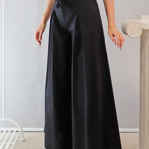 May include: Black wide-leg pants with a high waist and a matching fabric belt. The pants have a pleated front and a flowing silhouette. The model is wearing a patterned blouse and sandals. The pants are made of a smooth, opaque fabric.