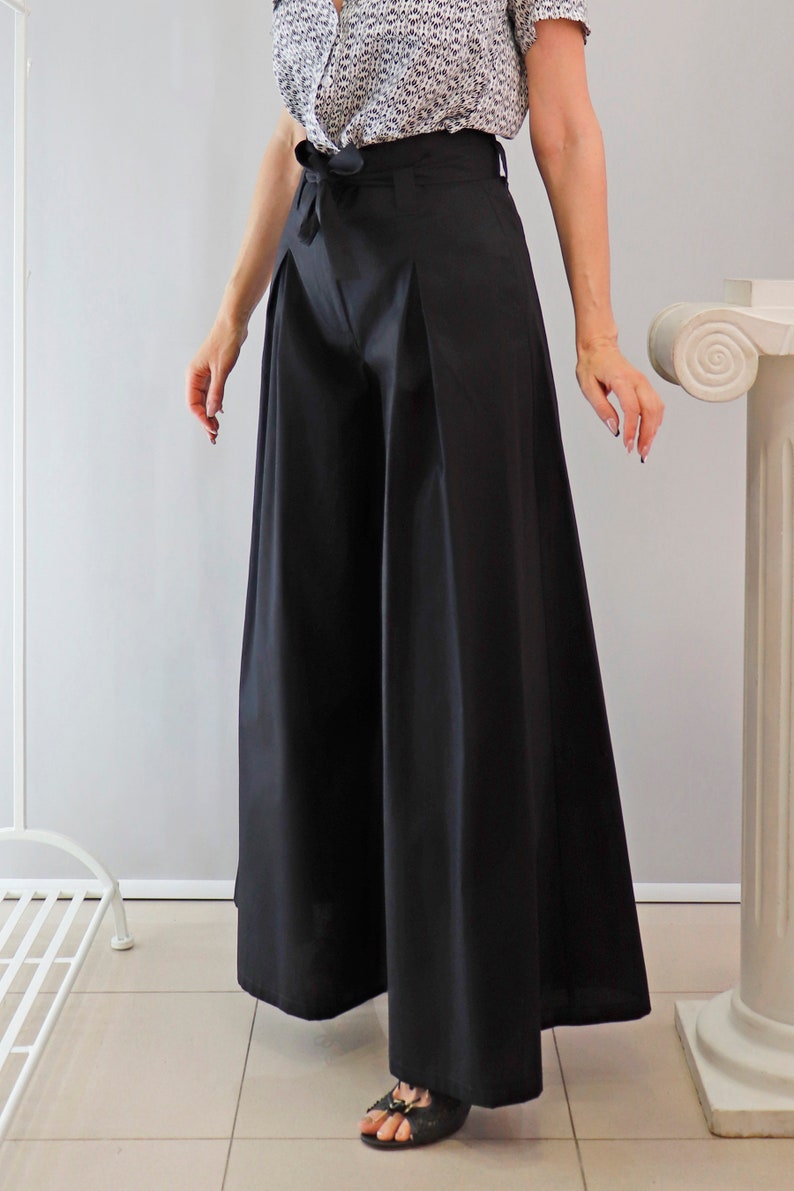 May include: Black wide-leg pants with a high waist and a matching fabric belt. The pants have a pleated front and a flowing silhouette. The model is wearing a patterned blouse and sandals. The pants are made of a smooth, opaque fabric.