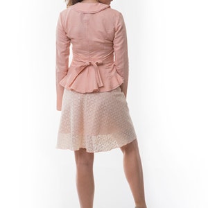 May include: A light pink long-sleeved jacket with a wide collar and a tie-back peplum waist, paired with a knee-length skirt featuring a delicate white lace overlay pattern. The ensemble is completed by beige wedge sandals with ankle straps.