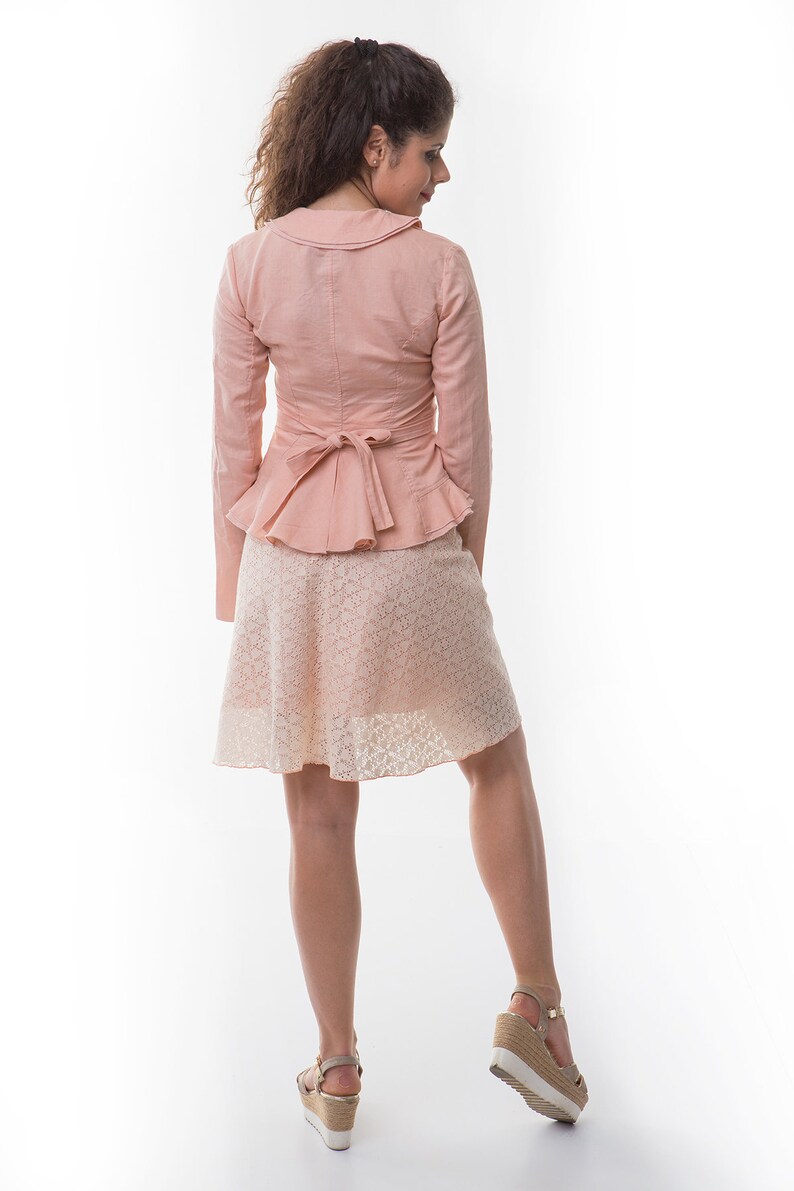 May include: A light pink long-sleeved jacket with a wide collar and a tie-back peplum waist, paired with a knee-length skirt featuring a delicate white lace overlay pattern. The ensemble is completed by beige wedge sandals with ankle straps.