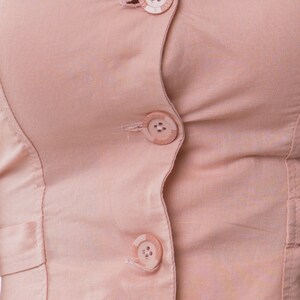 May include: A light pink button-down shirt featuring a ruffled collar and three circular buttons down the front. Each button is embossed with the text BONELLI. The garment is constructed from a solid-colored fabric with visible vertical seams.