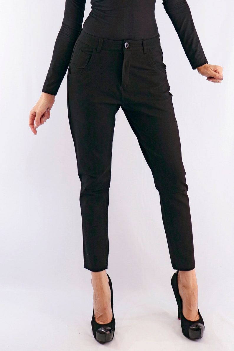 May include: Black trousers with a button closure and pockets. The pants are paired with black high heels. The outfit is all black, creating a sleek and streamlined look. The trousers appear to be made of a smooth fabric.