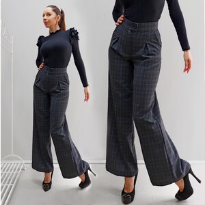May include: High-waisted wide-leg trousers in a dark gray plaid pattern featuring a structured waistband with a single button closure, front pleats, and side pockets, paired with a black long-sleeved top with ruffled shoulder detailing.