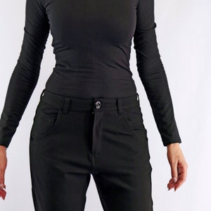 May include: Black long-sleeved top with a high neckline and lace detailing across the shoulders. Paired with black pants. The pants have front pockets and a button closure. The outfit is all black.