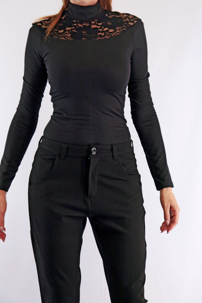 May include: Black long-sleeved top with a high neckline and lace detailing across the shoulders. Paired with black pants. The pants have front pockets and a button closure. The outfit is all black.