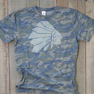 May include: A blue and green camouflage t-shirt with a white graphic of a Native American headdress.
