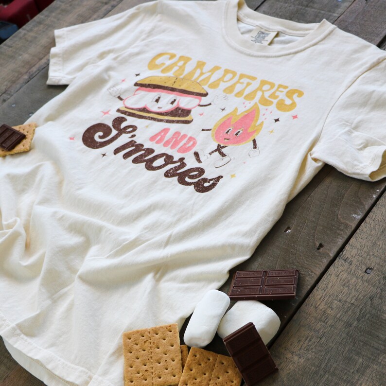 May include: A white t-shirt featuring a graphic of a smiling s'more and a fire with the text "Campfires and Smores". The shirt is surrounded by graham crackers, chocolate bars, and marshmallows.