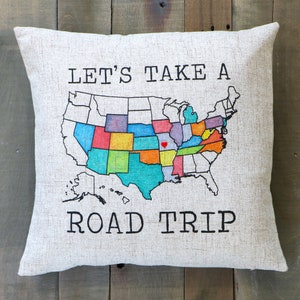 May include: A white throw pillow with the text "LET'S TAKE A ROAD TRIP" printed in black. A colorful map of the United States is printed in the center of the pillow with a red heart marking a location.