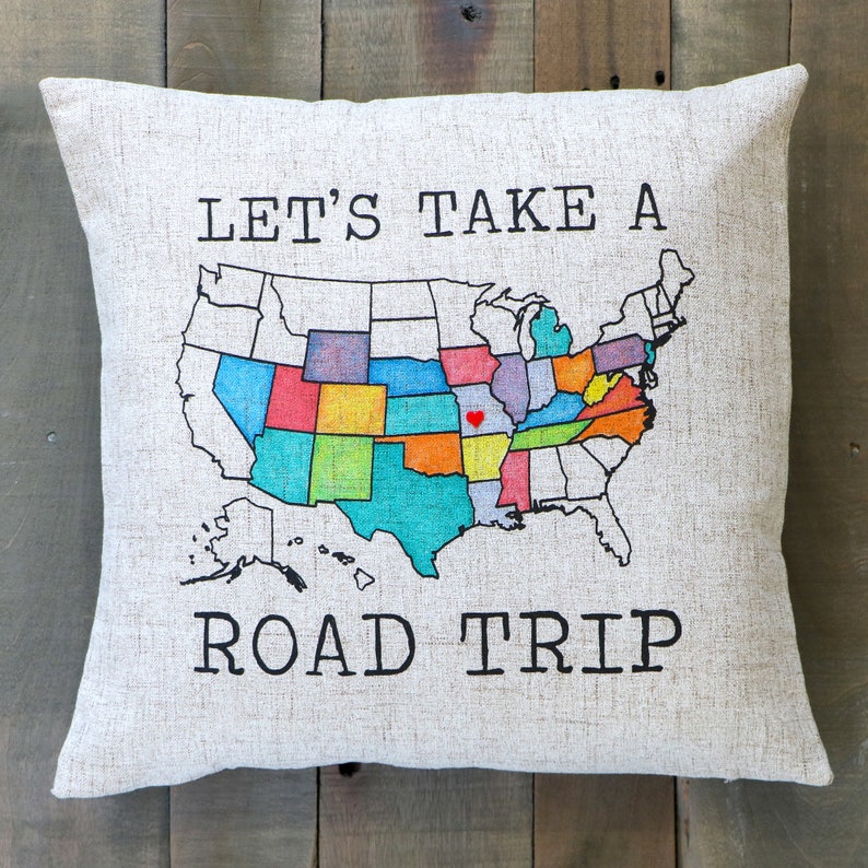 May include: A white throw pillow with the text "LET'S TAKE A ROAD TRIP" printed in black. A colorful map of the United States is printed in the center of the pillow with a red heart marking a location.