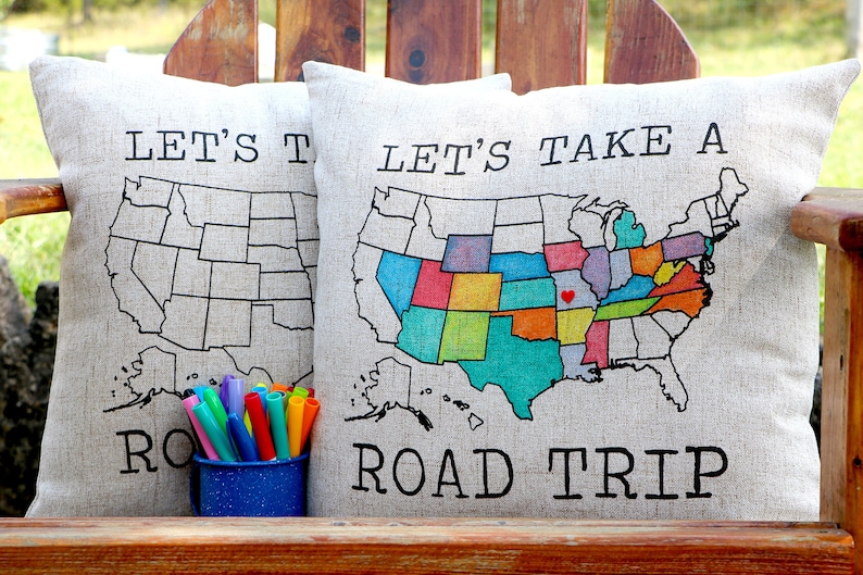 May include: Two beige throw pillows with the text "Let's Take A Road Trip" printed on them. The pillows feature a map of the United States with each state colored in a different color. One pillow has a blank map, while the other has a map with each state colored in. A set of colorful markers is in front of the pillows.