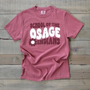 May include: A maroon t-shirt with a white graphic that says "School of the Osage Indians" with a white daisy with a smiley face.