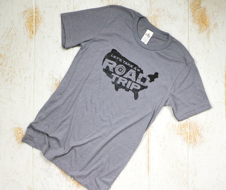 Op de afbeelding: Een grijs T-shirt met een zwarte afbeelding van de Verenigde Staten met de tekst "Let's Take a Road Trip" erop.