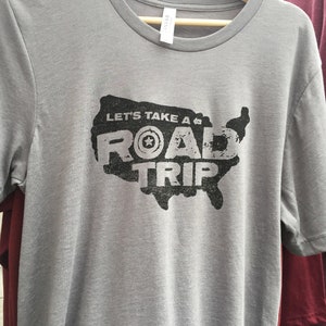 Op de afbeelding: Een grijs T-shirt met een zwarte afbeelding van een gestileerde kaart van de Verenigde Staten met de tekst "Let's Take a Road Trip" en een ster in een cirkel.