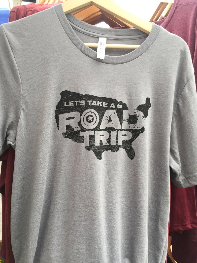Op de afbeelding: Een grijs T-shirt met een zwarte afbeelding van een gestileerde kaart van de Verenigde Staten met de tekst "Let's Take a Road Trip" en een ster in een cirkel.