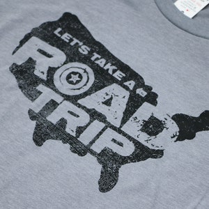 Op de afbeelding: Een grijs T-shirt met een zwart-witte afbeelding van de Verenigde Staten. De afbeelding zegt "Let's Take a Road Trip" met een gestileerde ster in een cirkel.
