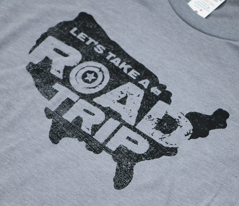 Op de afbeelding: Een grijs T-shirt met een zwart-witte afbeelding van de Verenigde Staten. De afbeelding zegt "Let's Take a Road Trip" met een gestileerde ster in een cirkel.