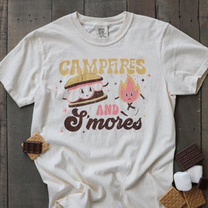 May include: A beige t-shirt featuring a cartoon s'more and a campfire. The text reads "Campfires and S'mores".