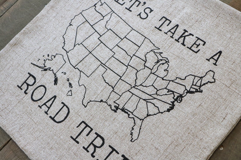 May include: A beige linen throw pillow with black text that reads "It's Take A Road Trip" and a black outline of the United States map with state borders.