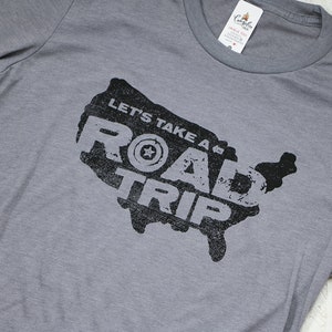 Op de afbeelding: Grijs T-shirt met een zwarte afbeelding van de kaart van de Verenigde Staten. De afbeelding bevat de tekst "Let's Take a Road Trip" en een gestileerde band met een ster in het midden.