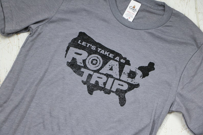 Op de afbeelding: Grijs T-shirt met een zwarte afbeelding van de kaart van de Verenigde Staten. De afbeelding bevat de tekst "Let's Take a Road Trip" en een gestileerde band met een ster in het midden.