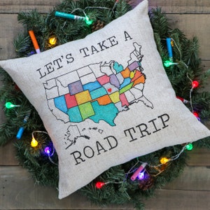 May include: A beige throw pillow with the text "Let's Take A Road Trip" printed in black. A colorful map of the United States is printed on the pillow, with a red heart marking a location.