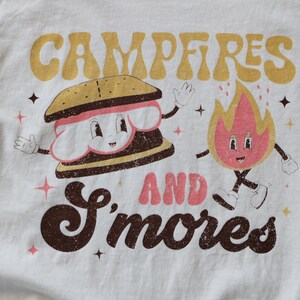 May include: A white t-shirt with a brown and yellow graphic that says "Campfires and S'mores". The graphic features a cartoon s'more and a cartoon fire.