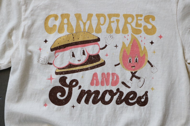 May include: A white t-shirt with a brown and yellow graphic that says "Campfires and S'mores". The graphic features a cartoon s'more and a cartoon fire.