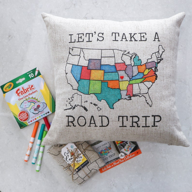 May include: A beige pillow with the text "Let's Take A Road Trip" printed on it. The pillow features a map of the United States with each state colored in a different color. A set of fabric markers and a small fabric bag with the text "Let's Take A Road Trip Collection" are also pictured.