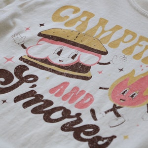 May include: A white t-shirt featuring a brown and pink graphic of a smiling s'more and a fire with the text "Campfire and Smores".