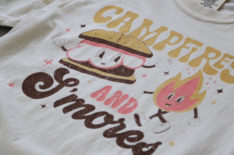 May include: A white t-shirt featuring a brown and pink graphic of a smiling s'more and a fire with the text "Campfire and Smores".