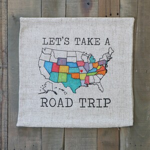 May include: A beige canvas pillow with a black and white graphic of a map of the United States with the text "Let's Take A Road Trip".