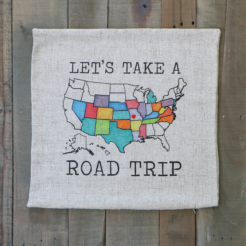May include: A beige canvas pillow with a black and white graphic of a map of the United States with the text "Let's Take A Road Trip".