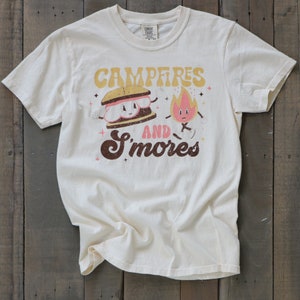 May include: A cream-coloured t-shirt featuring a graphic of a smiling s'more and a fire. The text reads "Campfires and S'mores" in a retro font.