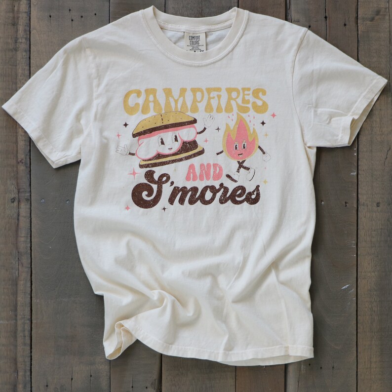 May include: A cream-coloured t-shirt featuring a graphic of a smiling s'more and a fire. The text reads "Campfires and S'mores" in a retro font.