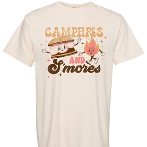 May include: A cream coloured t-shirt with a brown graphic of a smiling s'more and a smiling fire with the text "Campfires and S'mores".