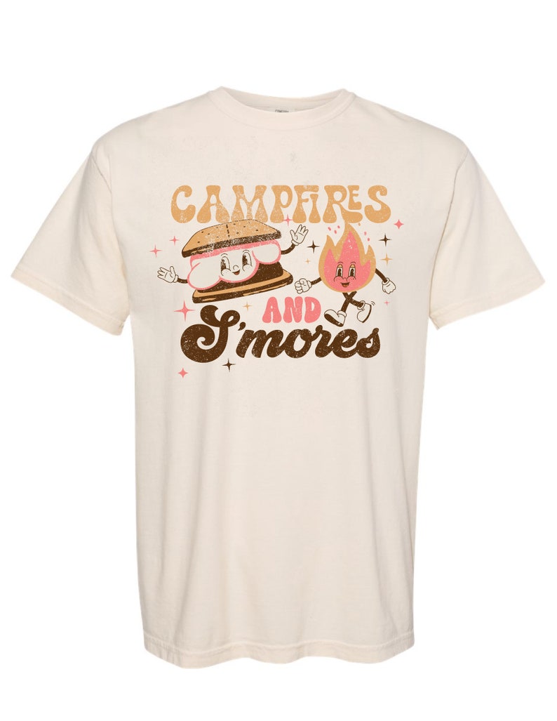 May include: A cream coloured t-shirt with a brown graphic of a smiling s'more and a smiling fire with the text "Campfires and S'mores".