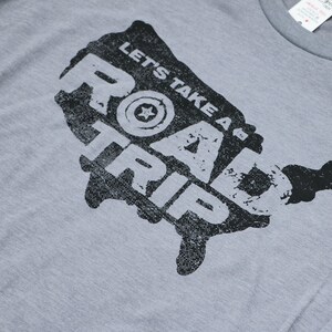 Op de afbeelding: Een grijs T-shirt met een zwarte afbeelding van de Verenigde Staten met de tekst "Let's Take A Road Trip" erop gedrukt.
