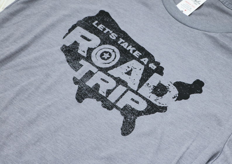 Op de afbeelding: Een grijs T-shirt met een zwarte afbeelding van de Verenigde Staten met de tekst "Let's Take A Road Trip" erop gedrukt.