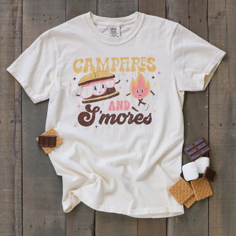 May include: A white t-shirt featuring a graphic of a smiling s'more and a fire with a face. The text "Campfires and S'mores" is printed in brown and black lettering.