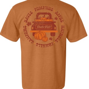 Puede incluir: Una camiseta marrón con un gráfico de estilo vintage de una camioneta llena de calabazas. El texto alrededor de la camioneta dice "Pumpkins, Apples, Hayrides, Campfires, Leaves, Flannels, Hello Fall!"