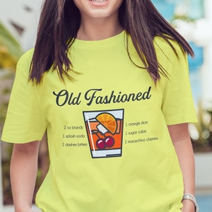 May include: A bright yellow t-shirt with a graphic of an Old Fashioned cocktail recipe. The text on the shirt reads "Old Fashioned" and includes the ingredients: 2 oz bourbon, 1 splash soda, 2 dashes bitters, 1 orange slice, 1 sugar cube, 1 maraschino cherry.