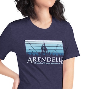 May include: A navy blue t-shirt with a graphic of a castle and mountains in a blue and white color scheme. The text "Arendelle" and "A land of frozen adventures" is printed on the shirt.