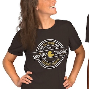 May include: Two people wearing black t-shirts with a white logo that says "Tap Room The Snuggly Duckling Karaoke Lounge" and a yellow rubber duck illustration.