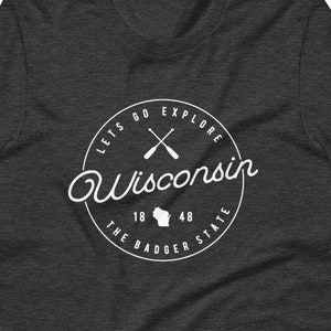 May include: A dark heather grey t-shirt with a white circle graphic featuring the text "Lets Go Explore Wisconsin", crossed paddles, the state outline, and "The Badger State 18 48".