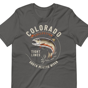 May include: A dark gray t-shirt with a circular design featuring a trout jumping out of the water. The design includes the text "Colorado Collection", "Tight Lines", "South Platte River", and a small red fishing lure.