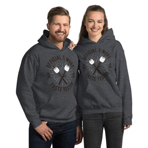 K&ouml;nnte beinhalten: Zwei Personen tragen dunkelgraue Kapuzenpullover. Die Kapuzenpullover haben einen Aufdruck mit der Aufschrift "OFFICIAL S'MORE TASTE TESTER" mit einem Design aus gekreuzten Marshmallow-R&ouml;stst&auml;ben. Die Kapuzenpullover haben eine Vordertasche und eine Kapuze.