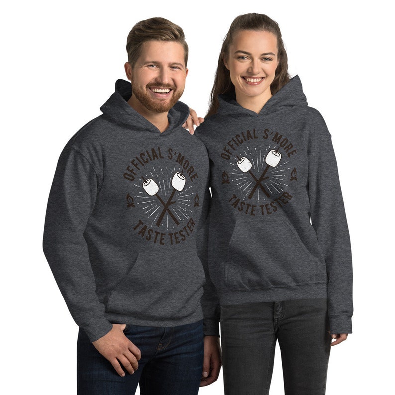 K&ouml;nnte beinhalten: Zwei Personen tragen dunkelgraue Kapuzenpullover. Die Kapuzenpullover haben einen Aufdruck mit der Aufschrift "OFFICIAL S'MORE TASTE TESTER" mit einem Design aus gekreuzten Marshmallow-R&ouml;stst&auml;ben. Die Kapuzenpullover haben eine Vordertasche und eine Kapuze.