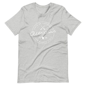 May include: A heather grey t-shirt with a white circle graphic that says "Let's Go Explore Wisconsin" with a white outline of the state of Wisconsin and two crossed paddles inside the circle. The text "The Badger State" is written below the state outline and the years "1848" are written above the state outline.